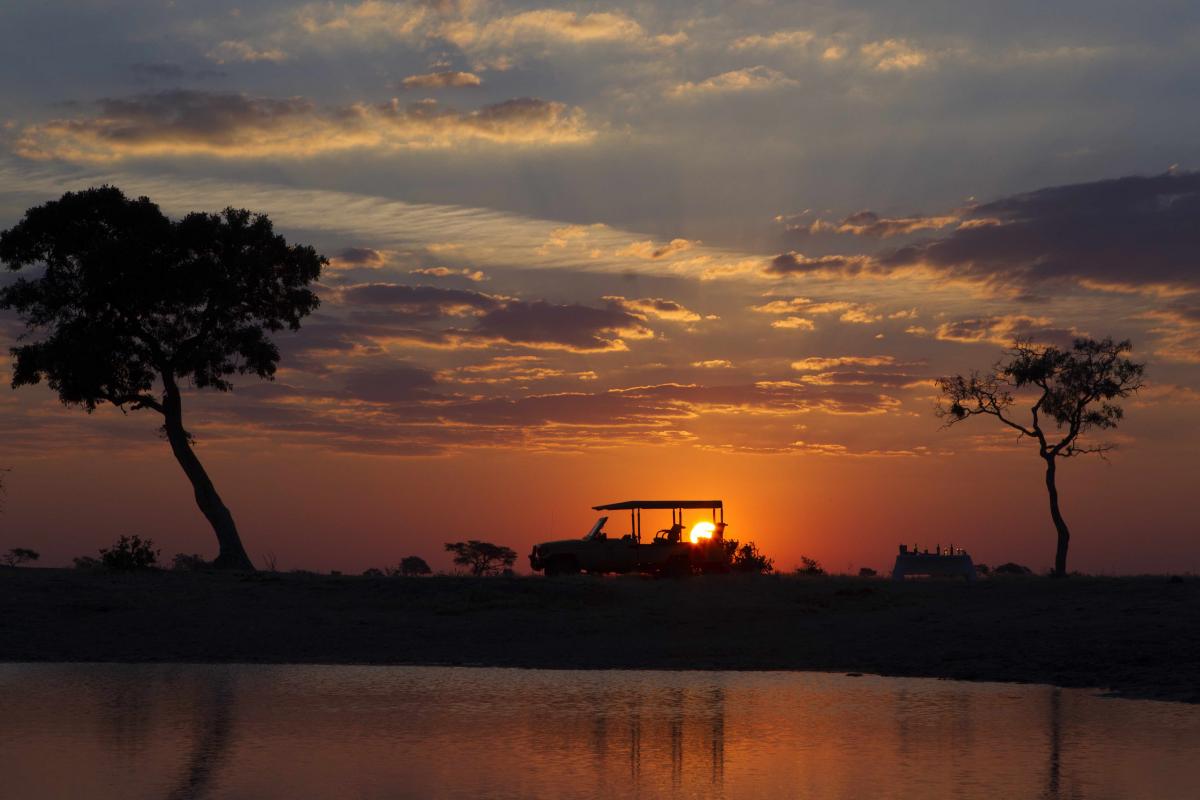 Namibi mit einem Sonnenuntergang und den Umrissen von einem Safari Jeep und einem Tisch mit Candlelight Dinner im Sonnenuntergang in seinen glühenden Farben