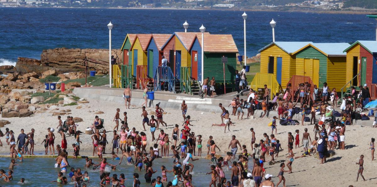 die bunten Häuschen dienen am Strand von Kapstadt als Umkleiden und sind als Fotomotiv sehr beliebt. Davor baden Einheimische und Touristen am weißen Sandstrand im blauen Meer. Die Sonne scheint